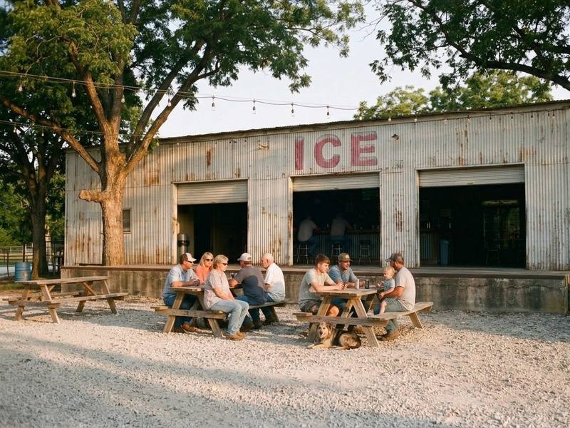 The tin icehouse in late afternoon light, three doors rolled up, the faded red ICE over the middle door, and a mixed crowd at picnic tables with a dog under one.